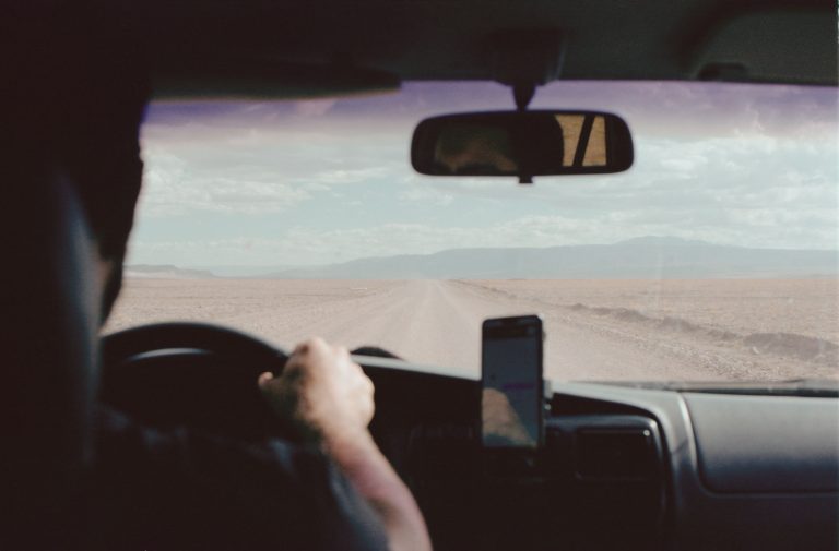 A driver's hand on a steering wheel viewed from inside a car on a desert road.